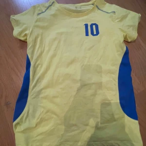 Gul fotbollströja med nummer 10 - Säljer en gul fotbollströja med blå detaljer och nummer 10 på både fram- och baksidan. Texten 'Sverige' är tryckt på ryggen. Tröjan har korta ärmar och blå paneler på sidorna.