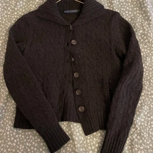 Stickad Ralph Lauren kofta - Gjord i 80% ull och använd några gången. L i ungdomsstorlek och passar mig XS-S. Svart/mörkgrå färg. Enda defekten är en knapp som fattas längst ner men går lätt att sy ny🪽