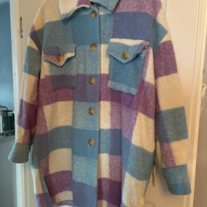 Rutig ulljacka från Italien - Säljer en mysig rutig jacka i ull med stora rutor i lila, blått och beige. Jackan har långa ärmar, knappar framtill och två bröstfickor med lock. Den har även fransar på baksidan för en unik touch. Perfekt för kyliga dagar!