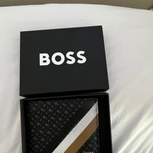 Plånbok från Boss - Snygg plånbok från Boss med ett stilrent mönster och färgblock i svart, vitt och brunt. Plånboken har flera kortfack och kommer i en elegant förpackning. Perfekt för att hålla ordning på dina kort med stil.