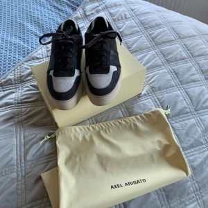 Svarta och vita sneakers från Axel Arigato - Snygga sneakers från Axel Arigato i svart och vitt. Skorna har en stilren design med snörning och en kontrasterande vit sula. Tillverkade i mocka och syntetmaterial, perfekta för en trendig look.