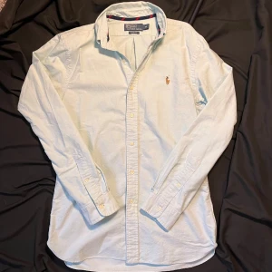 Ljusblå skjorta från Polo Ralph Lauren - Säljer en stilren ljusblå skjorta från Polo Ralph Lauren i slim fit. Skjortan har långa ärmar och klassiska knappar framtill. Den är broderad med det ikoniska Polo Ralph Lauren-märket på bröstet. Perfekt för en snygg och avslappnad look. Passar mig bra som är 178 kan passar större och mindre