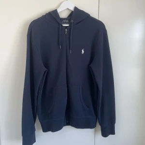 Mörkblå hoodie från Polo Ralph Lauren - Säljer en stilren mörkblå hoodie från Polo Ralph Lauren med dragkedja och det klassiska logotypbroderiet på bröstet. Storleken är S men passar M, själv är jag 185 och den sitter som en smäck. Inga defekter what so ever, mycket bra skick och sällan använd. Nypriset på tröjan är 2 295 kronor. 