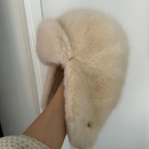 Vit pälsmössa från Barts - Säljer en supermjuk och fluffig beige/vit pälsmössa från Barts. Perfekt för att hålla värmen. Mössan har en klassisk design med öronlappar och ett justerbart band under hakan. Köpt för 799kr, använt 1v.