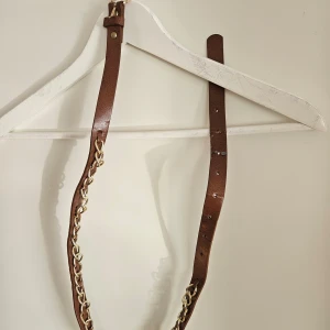 Brunt bälte med guldkedja - Snyggt brunt bälte med guldkedja som detalj. Perfekt för att ge en extra touch till din outfit. Justerbar längd med flera hål för perfekt passform.