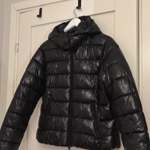Svart pufferjacka med huva - Säljer en svart pufferjacka med huva. Jackan har en glansig yta och dragkedja framtill. Perfekt för kyliga dagar.