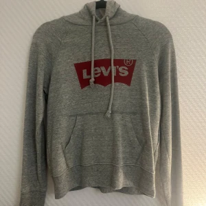 Grå hoodie från Levi's - Säljer en grå hoodie från Levi's i storlek XS. Den har en klassisk röd Levi's-logga på bröstet och en praktisk magficka. Perfekt för en avslappnad stil och passar bra till både höst och vår. Huvan har justerbara snören för extra komfort.