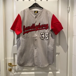 Grå och röd baseballtröja med Sundbyberg-tryck - Säljer en cool baseballtröja i grått med röda ärmar och Sundbyberg-tryck på bröstet. Nummer 35 pryder både fram- och baksidan. Perfekt för sportiga tillfällen eller som en avslappnad vardagslook. Knappar framtill för enkel på- och avtagning. Några sömmar som släppt m, se bild 3.