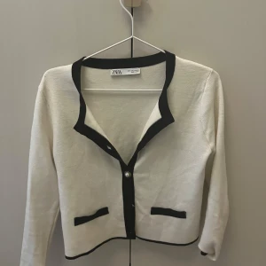 Vit kofta från Zara med svarta detaljer - Säljer en stilren vit kofta från Zara med svarta kanter och knappar. Den har en klassisk design med två dekorativa fickor framtill. Perfekt för både vardag och festliga tillfällen. 🖤🤍