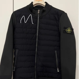 Svart cardiganjacka från Stone Island - Säljer en snygg svart jacka från Stone Island. Den har en quiltad design med dragkedja framtill och långa ärmar. Perfekt för höst och vinter, och den har den klassiska Stone Island-loggan på ärmen. Jackan är i grymt skick!