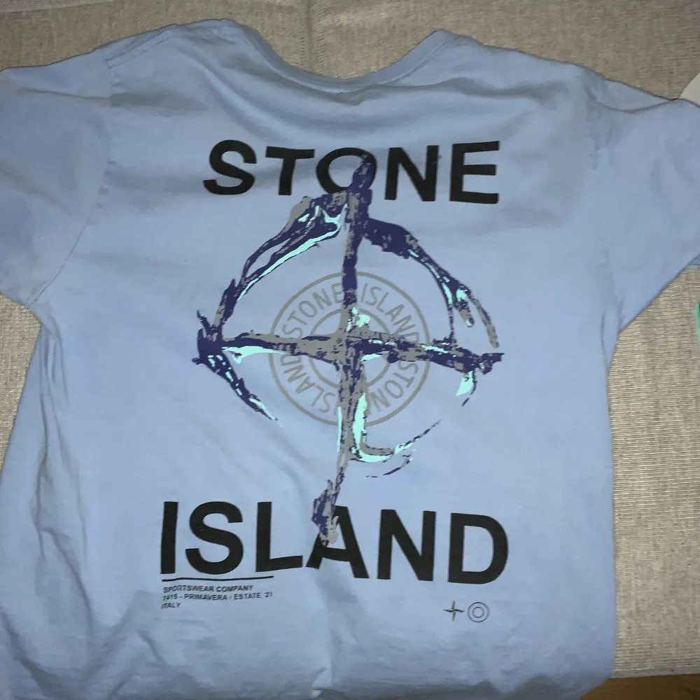 Säljer en snygg ljusblå t-shirt från Stone Island i 100% bomull. Den har en liten logga på framsidan och en stor, cool logga på baksidan. Perfekt för en avslappnad stil eller att bära till vardags. T-shirten är i bra skick och redo för nya äventyr!. T-shirts.