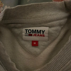 Grå sweatshirt från Tommy Jeans - Säljer en stilren grå sweatshirt från Tommy Jeans i storlek M. Tröjan har en klassisk design med ribbade muddar och en liten logga på bröstet. Perfekt för en avslappnad look eller som ett lager under jackan. Passar både höst och vår!