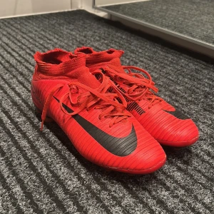 Röda Nike Mercurial fotbollsskor - Säljer ett par röda Nike Mercurial fotbollsskor i bra skick. De har en snygg svart Nike-logga och snörning för perfekt passform. Skorna har dobbar som ger bra grepp på planen. Perfekta för fotbollsträning eller match!