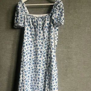 Blommig klänning i blått och vitt - Söt sommarklänning med blått blommönster på vit bakgrund. Klänningen har korta ärmar och en bekväm passform. Perfekt för en solig dag eller en avslappnad sommarfest. Materialet känns lätt och luftigt.