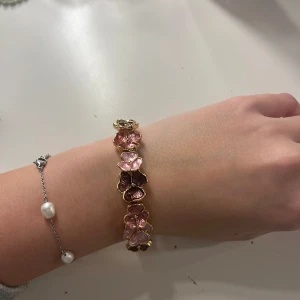 Guldarmband med rosa blommor - Säljer ett superfint guldarmband med rosa blommor. Armbandet har en elegant design med blommor i olika nyanser av rosa, perfekt för att ge en färgklick till din outfit. Det är elastiskt och passar de flesta handleder. Perfekt för både vardag och fest! 🌸