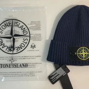 Stone Island mössa - Säljer en snygg blå ribbad mössa från Stone Island. Den har en broderad logga framtill med knappar på sidorna. Perfekt för att hålla sig varm och stilren under kalla dagar. Kommer i originalförpackning1.1