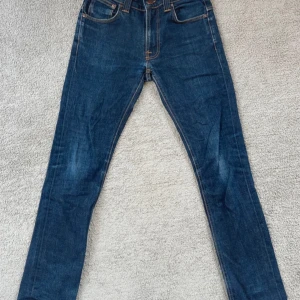 Blå jeans från Nudie Jeans - Säljer ett par snygga blå jeans från Nudie Jeans i mycket bra skick. De har en klassisk femficksdesign med orange sömmar och en knappgylf. Perfekta för både vardag och fest!