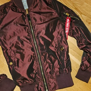 Vinröd bomberjacka från Alpha Industries - Säljer en snygg vinröd bomberjacka från Alpha Industries. Den har en klassisk design med dragkedja och ribbade muddar. Jackan är i mycket bra skick och perfekt för höst och vår. Den har en cool detalj med en röd tagg på ärmen. Passar perfekt för en casual look!