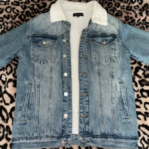 Jeansjacka med teddyfoder från Frilivin - Säljer en snygg jeansjacka från Frilivin i storlek M. Den har ett mysigt vitt teddyfoder och klassisk jeansblå färg. Jackan har knappar framtill och två bröstfickor med lock. Perfekt för höst och vår när du vill hålla stilen och värmen! 🧥
