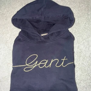 Svart hoodie från Gant - Säljer en snygg svart hoodie från Gant i storlek S. Den har en stor Gant-logga i guld på framsidan och en praktisk magficka. Perfekt för höst och vinter, och den är superbekväm att ha på sig. Huvan är stor och mysig, perfekt för kyliga dagar. 🖤✨