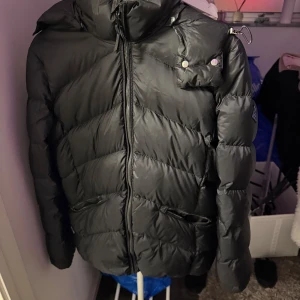 Svart pufferjacka från GANT - Säljer en svart pufferjacka från GANT i storlek medium. Jackan har en dragkedja framtill och en hög krage för extra värme. Perfekt för kalla vinterdagar. Den är i bra skick och redo för nya äventyr!