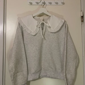 Tjocktröja - Säljer en superfin grå tröja från H&M med en stor vit volangkrage och knytband. Perfekt för en söt och stilren look. Tröjan är oanvänd🩶