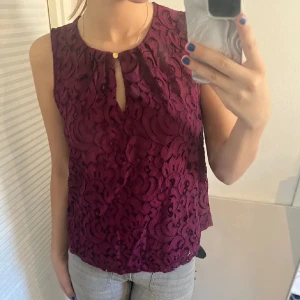 Vinröd spetsblus - Säljer en elegant vinröd spetsblus med ett vackert mönster. Blusen är ärmlös och har en liten knappdetalj vid halsen. Perfekt för både fest och vardag! 💃