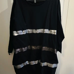 Svart klänning med silverdetaljer från Rainbow - Snygg svart klänning från Rainbow i storlek L. Den har trekvartsärmar och coola silverfärgade ränder som ger en glittrig touch. Perfekt för en festlig kväll eller en utekväll med vänner. Klänningen är i bra skick och redo att bäras!