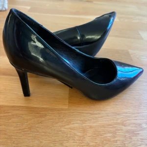 Svarta lackpumps med klack - Snygga svarta pumps i mycket bra skick. De har en elegant spetsig tå och en smal klack som ger en stilren look. Perfekta för fest eller en utekväll. Materialet är glansig lack och ger en lyxig känsla. Passar till både klänning och byxor!