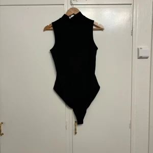 Svart ärmlös bodysuit - Snygg svart ärmlös bodysuit i ribbat material. Perfekt för en stilren look, oavsett om det är till vardags eller fest. Den har en hög hals och är superbekväm att bära. Passar perfekt under en kavaj eller med ett par jeans för en chic outfit.