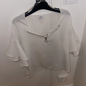 Vit blus med dragkedja från Linneox - Säljer en stilren vit topp från Linneox i storlek M. Den har en cool dragkedja framtill och volangdetaljer på ärmarna. Perfekt för både vardag och fest! 🕊️