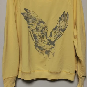Gul tröja från Hilfiger Denim - Säljer en cool gul tröja från Hilfiger Denim med ett stort tryck av en örn på framsidan. Tröjan har långa ärmar och är perfekt för en avslappnad stil. Den är i bra skick och passar perfekt till både jeans och shorts. Perfekt för vår och sommar!