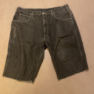 Svarta jeansshorts från Sean John - Säljer ett par svarta jeansshorts från Sean John. De har en avslappnad passform med råa kanter och en snygg broderad logga på framsidan. Perfekta för en casual look!