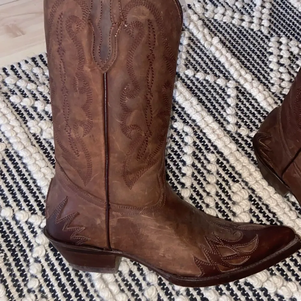 Säljer ett par snygga bruna cowboy boots i skinn. De har ett klassiskt broderat mönster och spetsig tå. Perfekta för en cool och avslappnad stil. Använd dem till både vardags och fest!. Skor.