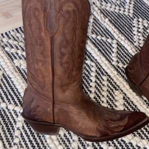 Säljer ett par snygga bruna cowboy boots i skinn. De har ett klassiskt broderat mönster och spetsig tå. Perfekta för en cool och avslappnad stil. Använd dem till både vardags och fest!