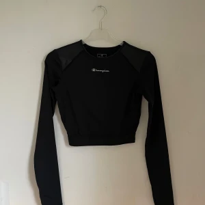 Svart croppad tröja från Champion - Säljer en snygg svart croppad tröja från Champion i storlek XS. Tröjan har långa ärmar och är perfekt för träning eller en casual look. Den är gjord i ett syntetmaterial som känns bekvämt mot huden. Champion-loggan finns både fram och bak, vilket ger en sportig touch.