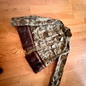 Camouflagejacka från Zara Woman - Säljer en cool camouflagejacka från Zara Woman. Den har långa ärmar och är dekorerad med bruna pälsdetaljer vid ärmsluten. Jackan har en klassisk krage och knappar framtill. Perfekt för en avslappnad och trendig look! Jättebra skick