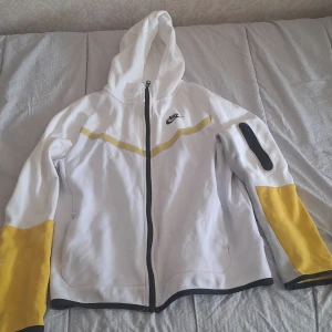 Vit och gul jacka från Nike - Säljer en vit och gul jacka från Nike i storlek XL. Jackan har en huva och dragkedja framtill. Den är vit med gula detaljer på ärmarna och en gul rand över bröstet. Det finns en ficka med dragkedja på ena ärmen. Jackan är i välanvänt skick med några fläckar på den gula delen av ärmen.