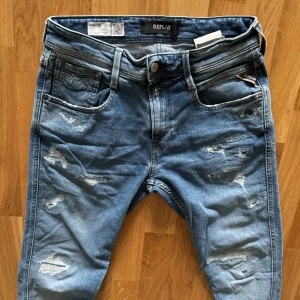 Blå jeans från Replay - Säljer ett par snygga blå jeans från Replay, modell Anbass. De har en cool slitningseffekt på framsidan och en klassisk femficksdesign. Jeansen är i en slim passform och har en knappgylf. Perfekta för en avslappnad men stilren look!