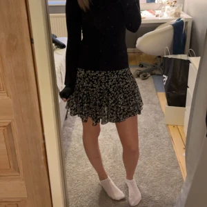 Svart blommig kjol - Säljer en superfin svart kjol med vita blommor. Den är kort och perfekt för både vardag och fest. Kjolen har en volangdetalj som ger den en extra söt touch. Materialet känns lätt och luftigt, vilket gör den perfekt för vår och sommar.