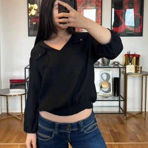Svart blus med v-ringning Zara  - Säljer en stilren svart blus med v-ringning och trekvartsärmar. Blusen har en avslappnad passform och är perfekt för både vardag och fest. Den har en liten knappdetalj på axeln och elastisk nederkant för en bekväm passform.