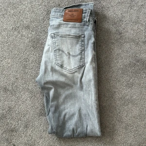 Grå jeans från Jack & Jones - Säljer ett par snygga grå jeans från Jack & Jones i modellen Slim/Glenn. De har en slim passform och är perfekta för en stilren look. Jeansen är i bra skick och har en klassisk femficksdesign med en läderpatch på baksidan. Perfekta för både vardag och fest!
