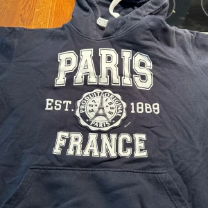 Mörkblå hoodie med Paris-tryck - Säljer en snygg och bekväm mörkblå hoodie med stort tryck av 'PARIS FRANCE' på framsidan. Hoodien har en klassisk känguruficka och justerbar huva med vita snören. Perfekt för en avslappnad stil eller en kylig dag ute. Passar både till vardags och för en casual look.