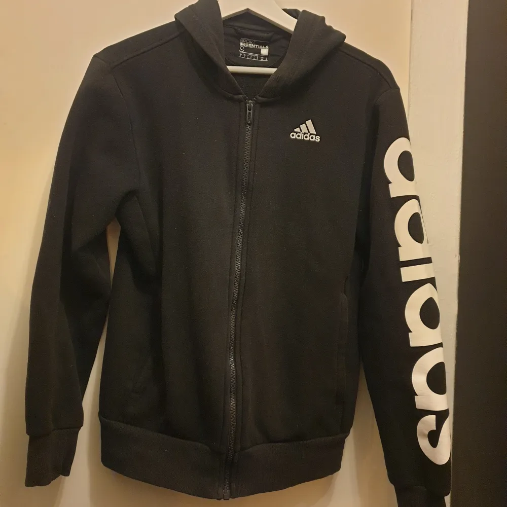 Säljer en svart hoodie från Adidas med dragkedja och deras klassiska logga på bröstet. Den har långa ärmar och Adidas tryckt längs ena ärmen. Perfekt för en sportig look eller bara för att chilla i. Superbekväm och stilren! Storlek S men passae M oxå. Hoodies.