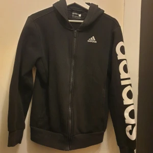 Svart hoodie från Adidas - Säljer en svart hoodie från Adidas med dragkedja och deras klassiska logga på bröstet. Den har långa ärmar och Adidas tryckt längs ena ärmen. Perfekt för en sportig look eller bara för att chilla i. Superbekväm och stilren! Storlek S men passae M oxå