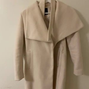 Beige kappa med bred krage - Elegant beige kappa med en bred och stilren krage. Kappan har långa ärmar och en rak passform, perfekt för kyligare dagar. Den har diskreta fickor på sidorna och en enkel design som ger ett sofistikerat intryck.