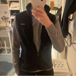 Mörkblå fleeceväst från Patagonia - Säljer en stilren grå fleeceväst från Patagonia. Västen har en dragkedja framtill och två praktiska fickor med dragkedjor. Perfekt för lager-på-lager under kyligare dagar. Patagonia-loggan är diskret placerad på bröstet.