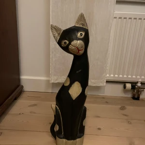 Dekorativ träskulptur av katt - Charmig träskulptur av en katt med svart och vit färg. Katten har en lång, elegant kropp och ett uttrycksfullt ansikte med stora ögon och ett rött leende. Perfekt som inredningsdetalj för kattälskare. Höjd 62cm. Finns totalt 4 st i olika storlekar 