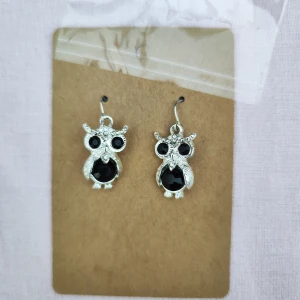 Silvriga uggleörhängen med svarta stenar - Charmiga örhängen i form av ugglor med detaljer i silver och svarta stenar. Perfekta för att ge en unik touch till din stil. Ugglorna har stora, svarta ögon och en rund svart sten på kroppen.