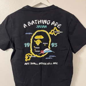 Svart t-shirt från A Bathing Ape - Säljer en svart t-shirt från A Bathing Ape med färgglatt tryck av Japan-karta och texten 'Ape Shall Never Kill Ape'. T-shirten har korta ärmar och en rund halsringning. Perfekt för streetwear-entusiaster! Size M